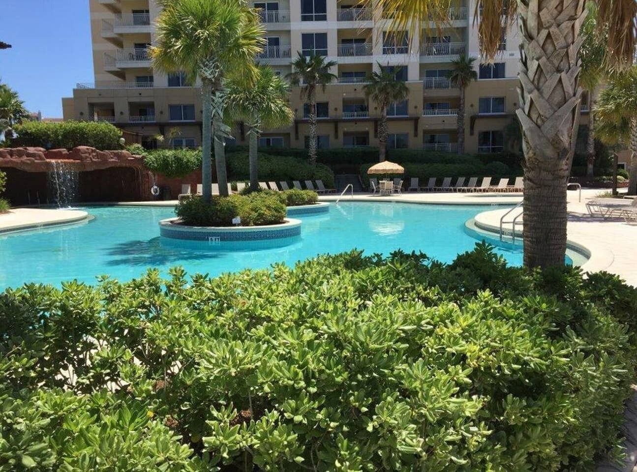 Una piscina rodeada de exuberante vegetación y palmeras, con un edificio de varios pisos al fondo.