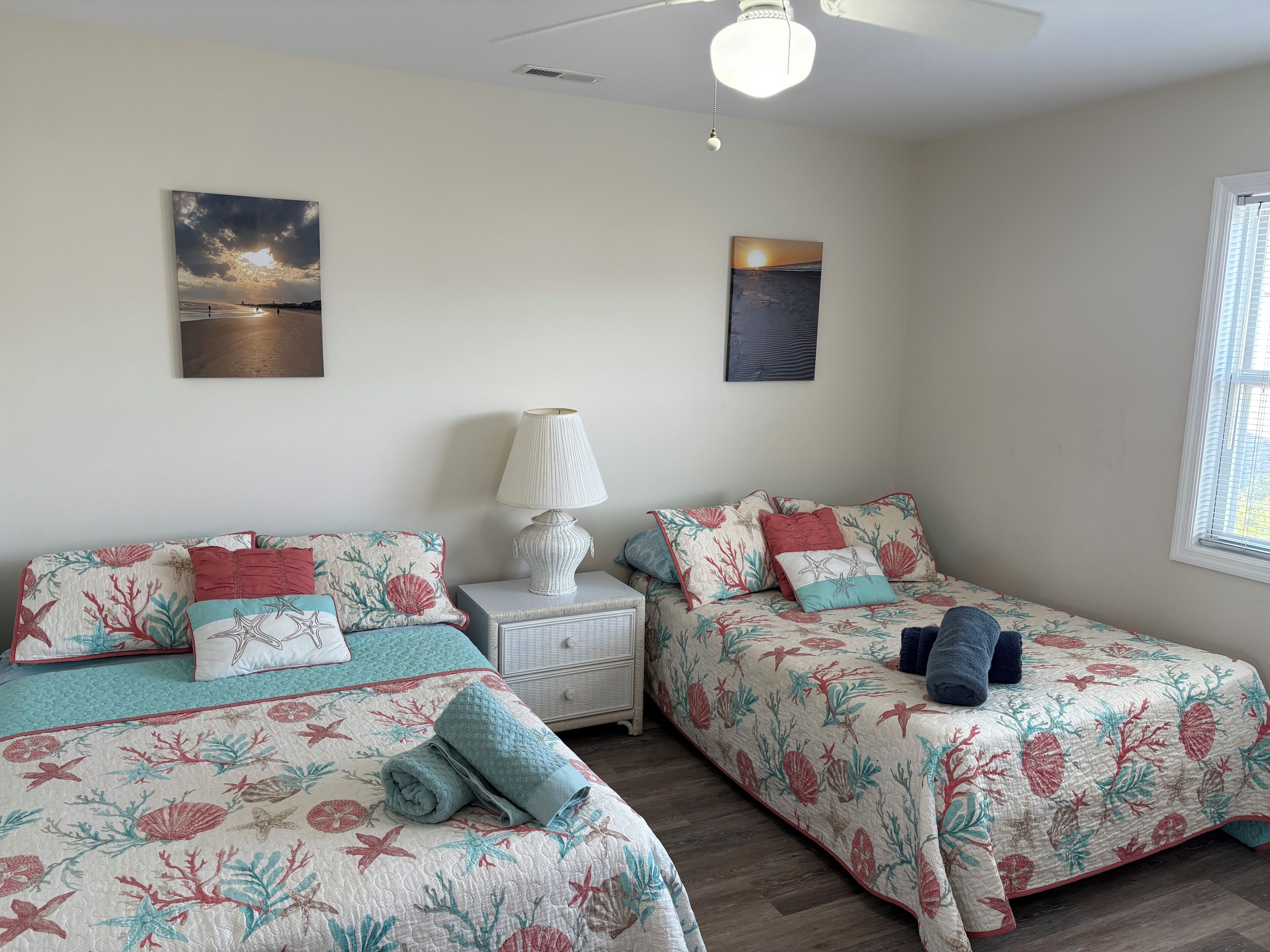 A bedroom with two beds, each with floral bedding, a bedside table with a lamp, and artwork on the walls.