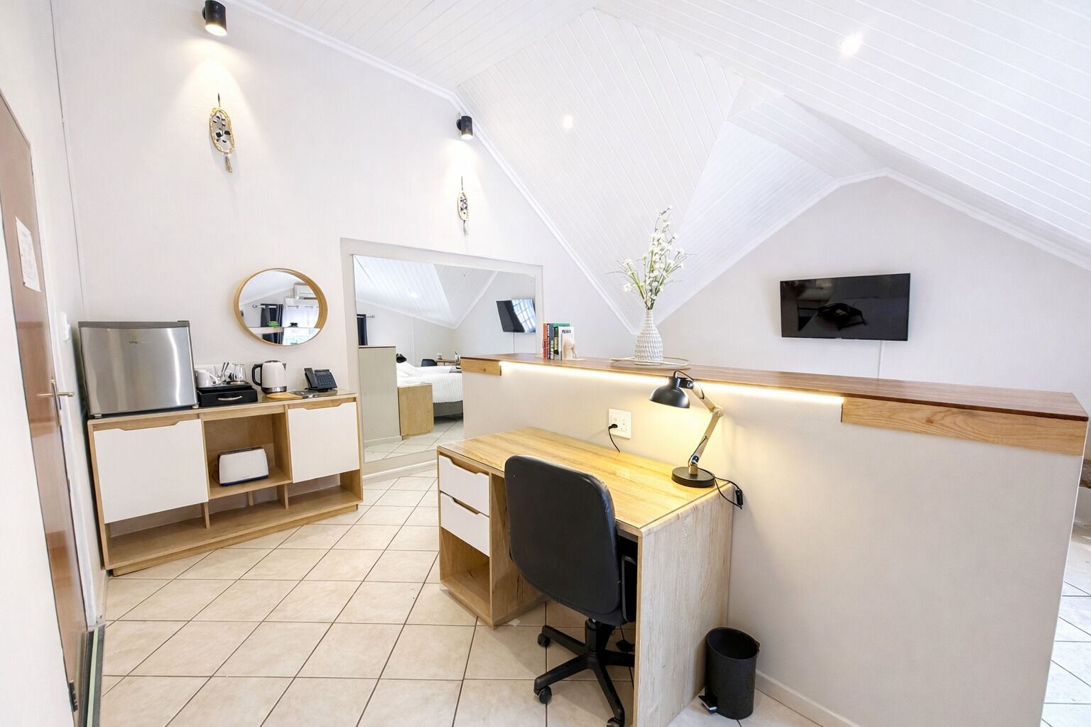 A modern, well-lit room with a wooden desk, a chair, a mirror, a small kitchenette, and a bed in the background.
