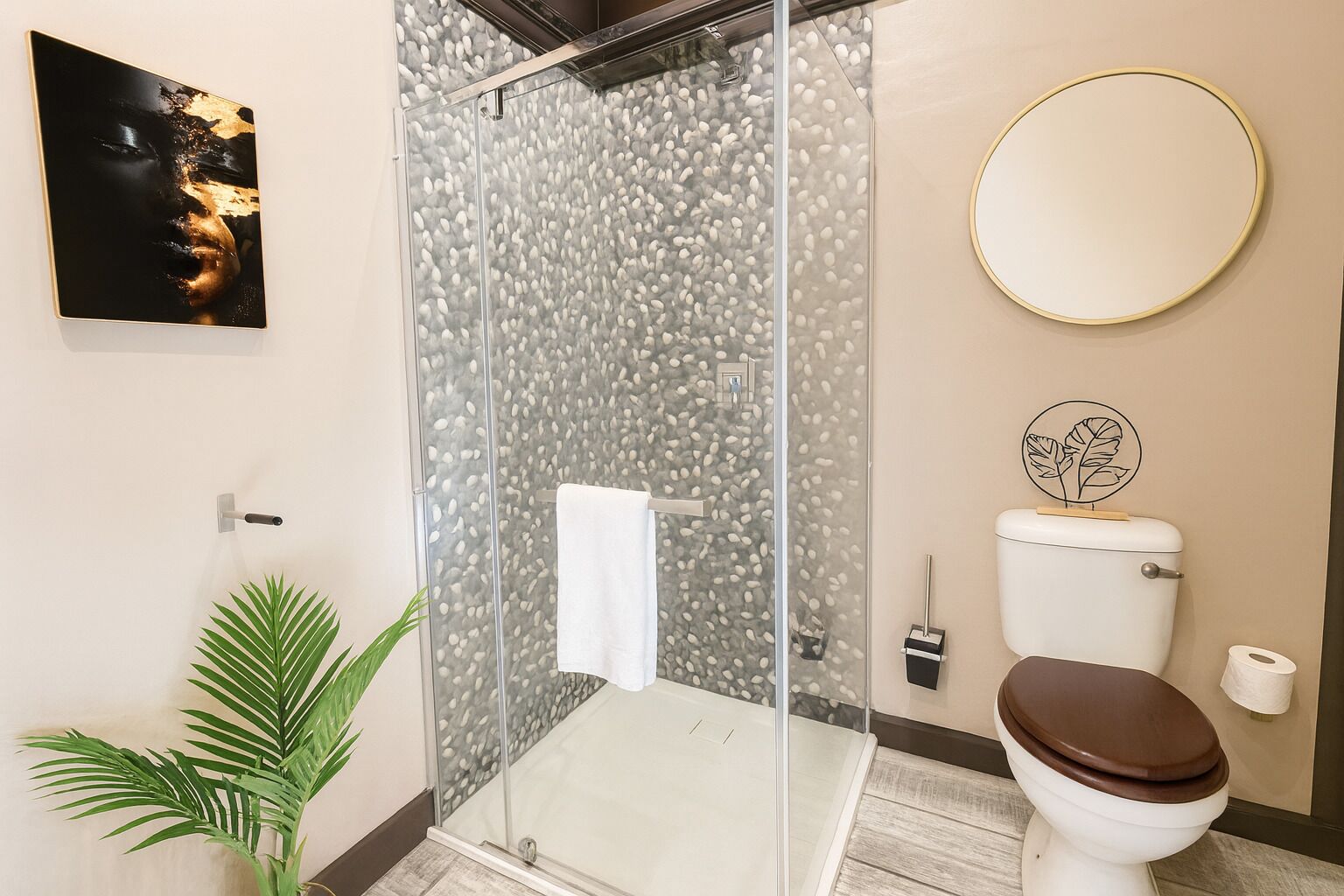 A modern bathroom with a glass shower enclosure, a toilet with a decorative lid, a round mirror, and a potted plant.