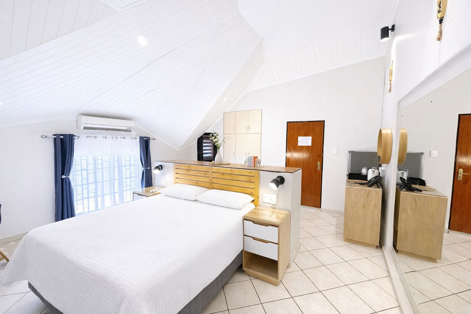Habitación de hotel con una cama grande, cabecera de madera, mesita de noche, espejo, armario y televisión.