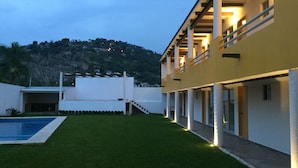 A modern building with a swimming pool, a well-maintained lawn, and a hillside in the background.