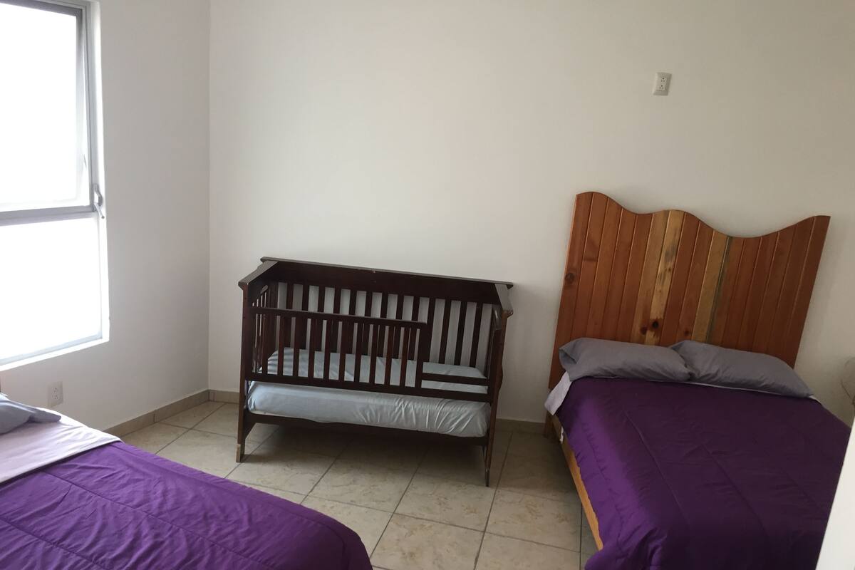 Una habitación con una cuna, una cama con cabecera de madera y una ventana.