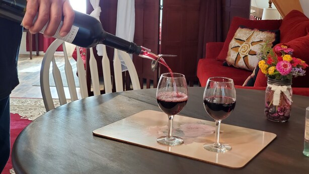 Una persona sirve vino en dos copas sobre una mesa en una habitación con un sofá rojo y un florero.
