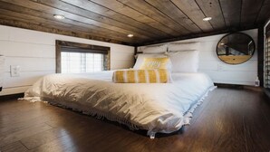 Una camera da letto con soffitto in legno, un letto grande con biancheria da letto bianca e un cuscino giallo, uno specchio rotondo a parete e una finestra con persiane.