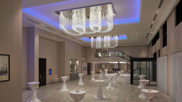 A spacious hotel lobby with multiple round tables, a grand chandelier, and a staircase leading to an upper level.