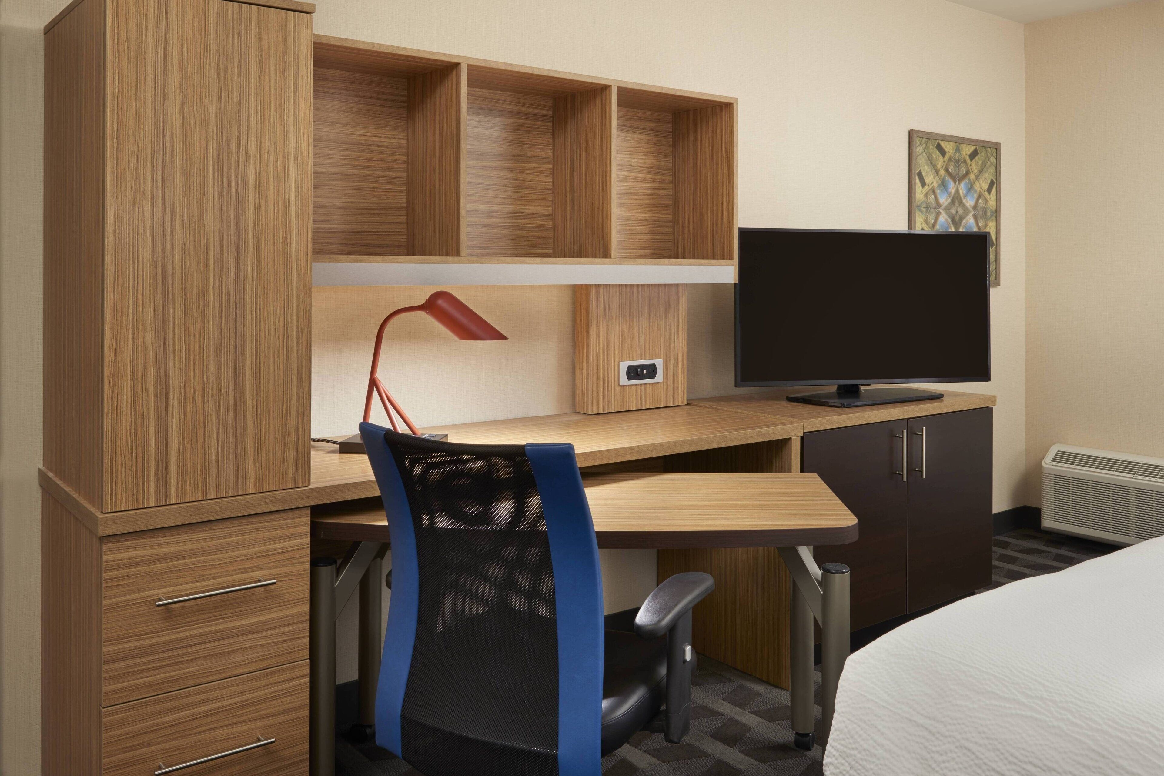 A hotel room with a wooden desk, a chair, a television, and a bed.