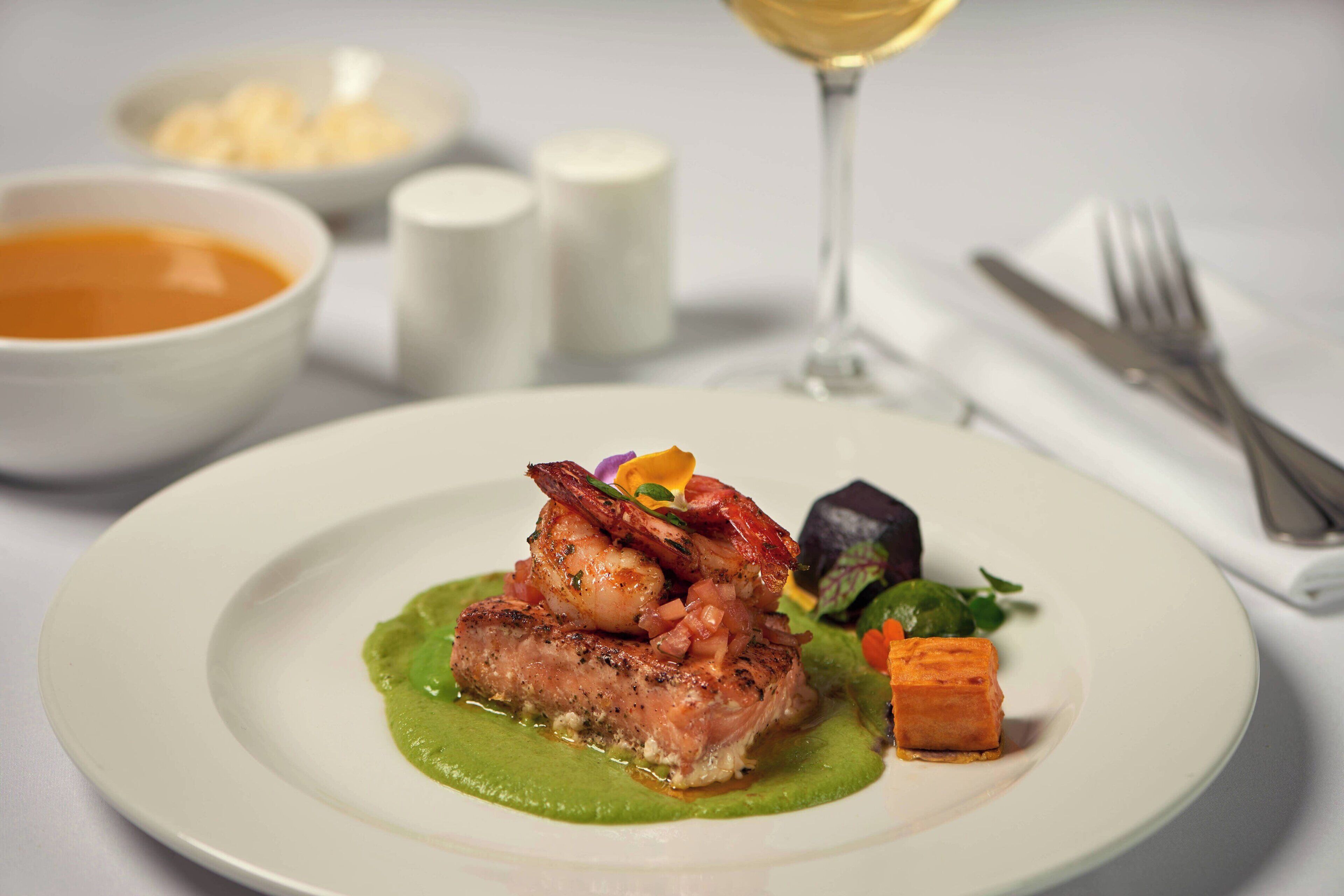 Un plato de comida acompañado de una copa de vino blanco, saleros, pimenteros, un tenedor y un cuchillo sobre una servilleta.