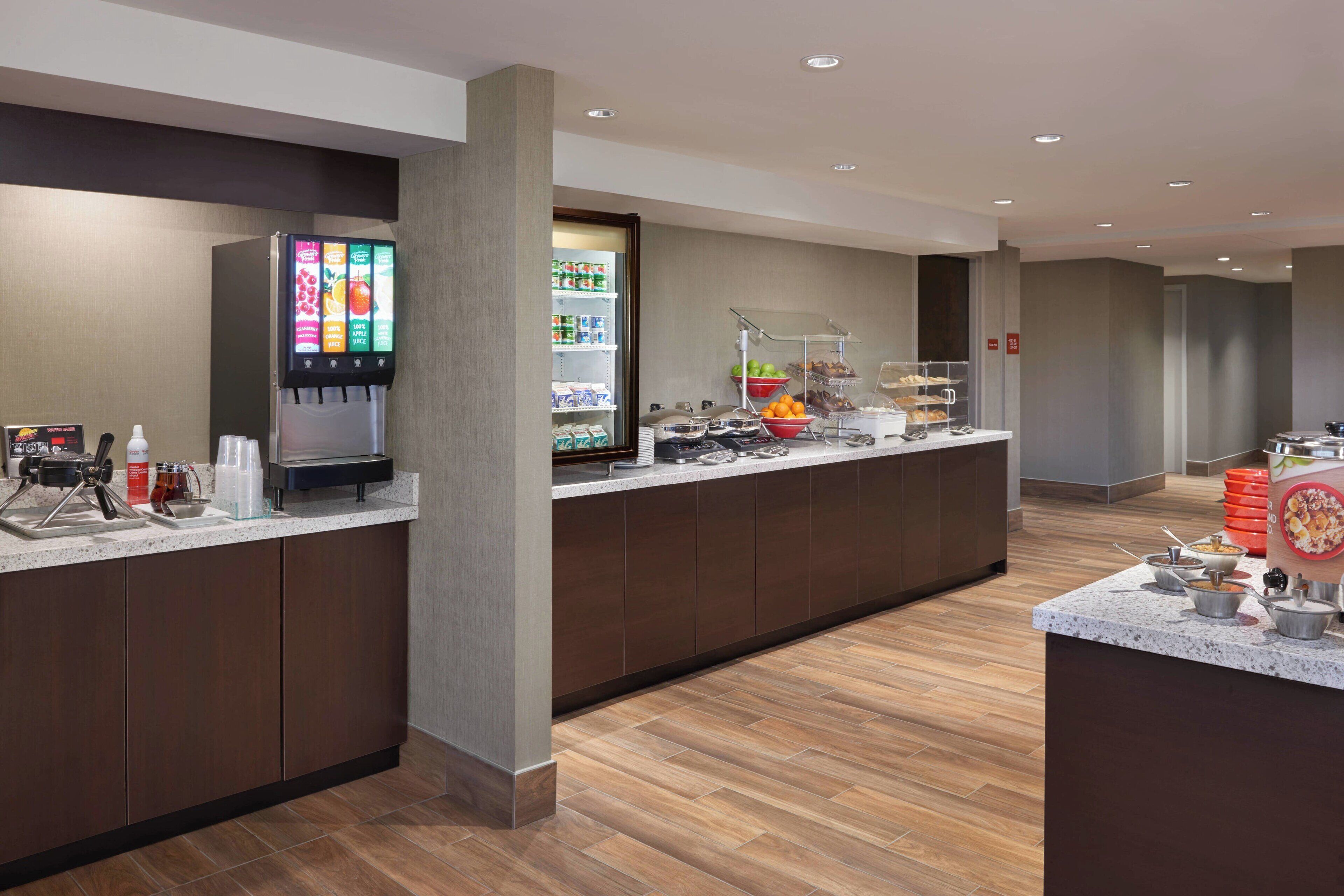 Un área de desayuno moderno en un hotel, con una estación autoservicio de bebidas, un refrigerador exhibidor y una variedad de opciones gastronómicas.