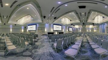 A conference room with rows of chairs, a carpeted floor, and a high ceiling with recessed lighting and chandeliers.