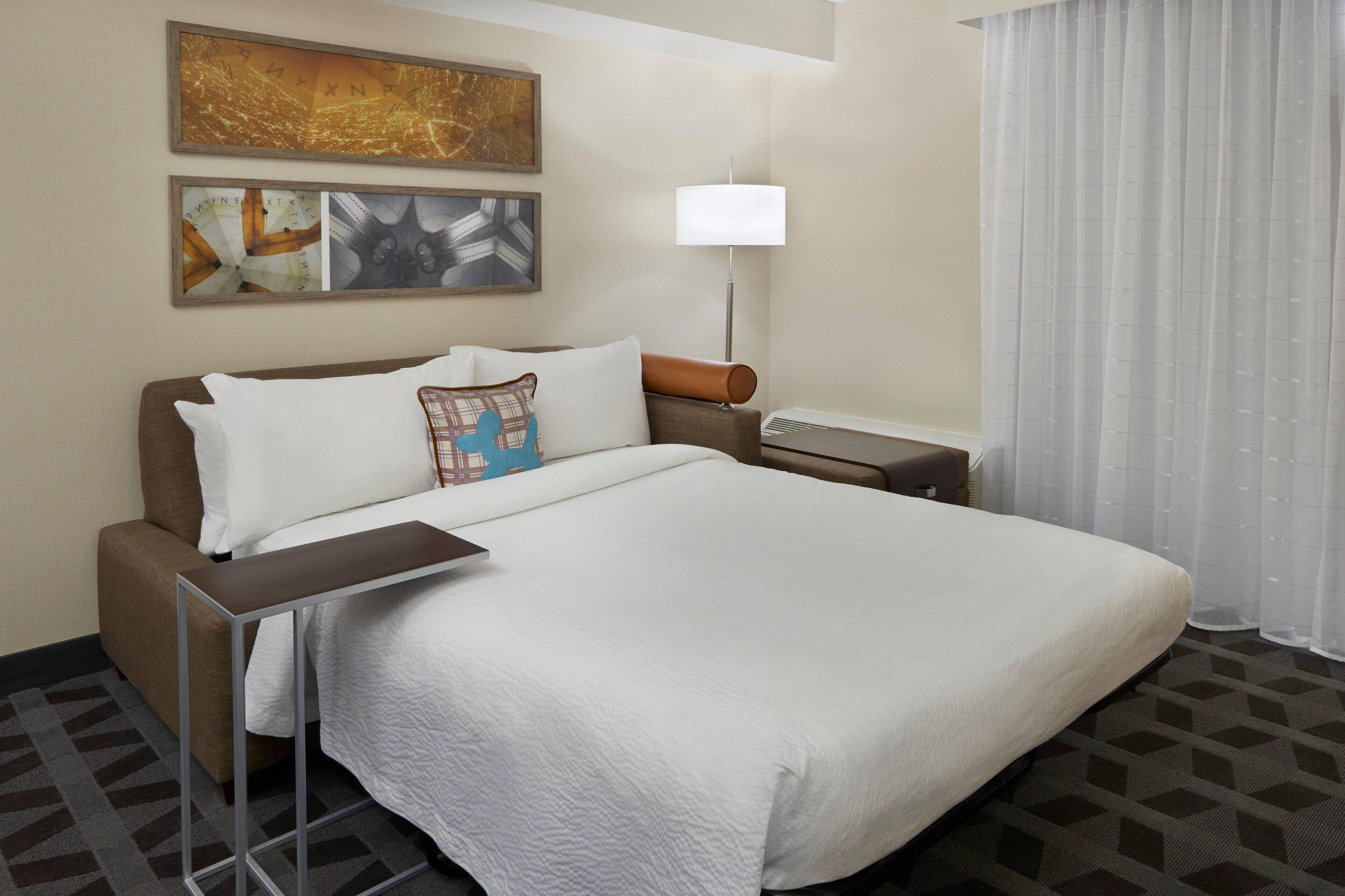A hotel room with a large bed, a bedside table, a lamp, and a view of the outside through sheer curtains.