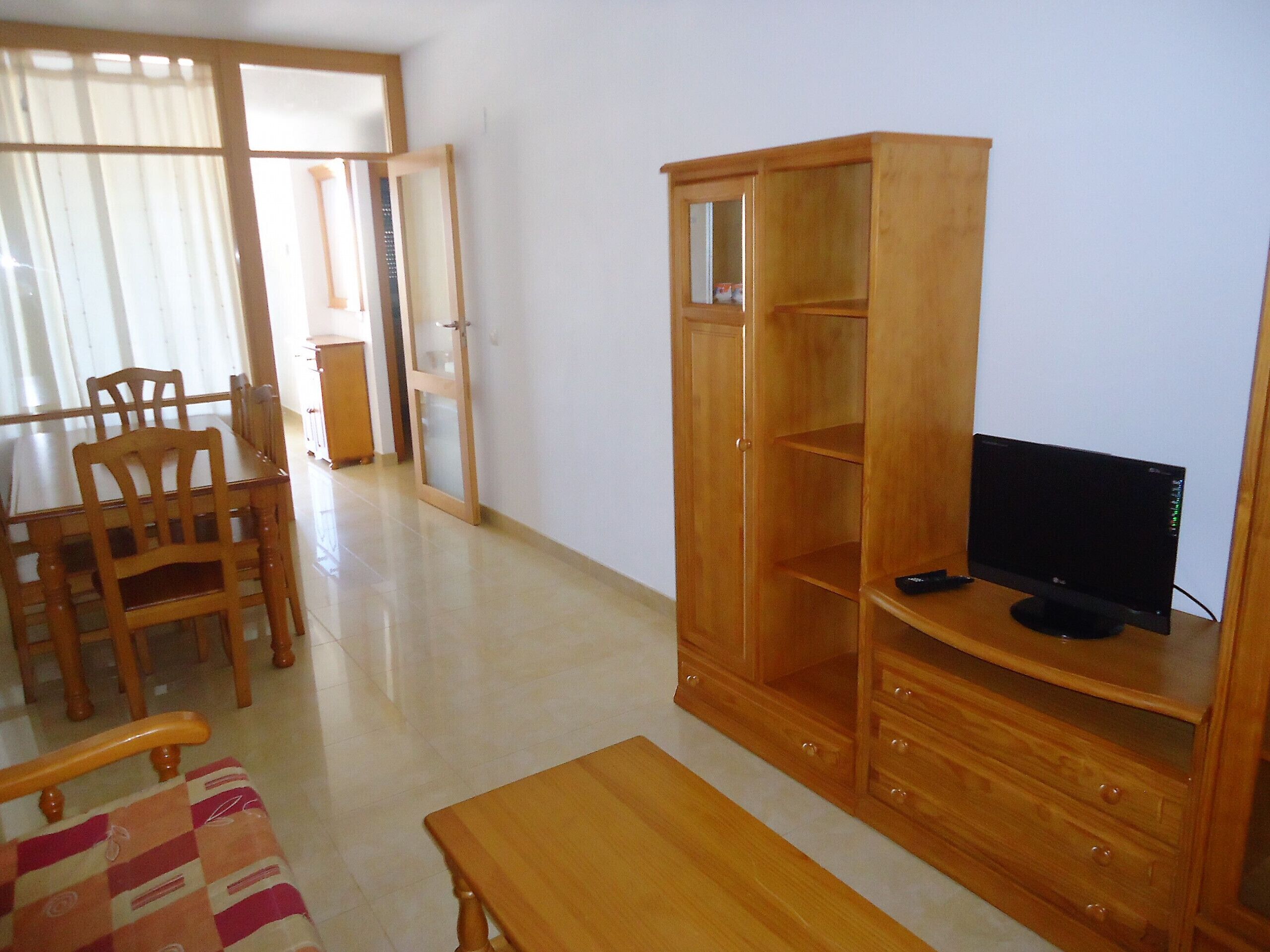 Una sala de estar con un mueble bajo de madera para el televisor, un televisor, una mesa de comedor de madera con sillas y un ventanal corredero que da a otra habitación.