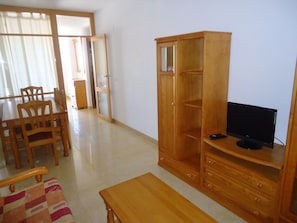Un salon comprenant un meuble TV en bois, un téléviseur, une table à manger en bois avec des chaises, et une porte-fenêtre coulissante donnant sur une autre pièce.