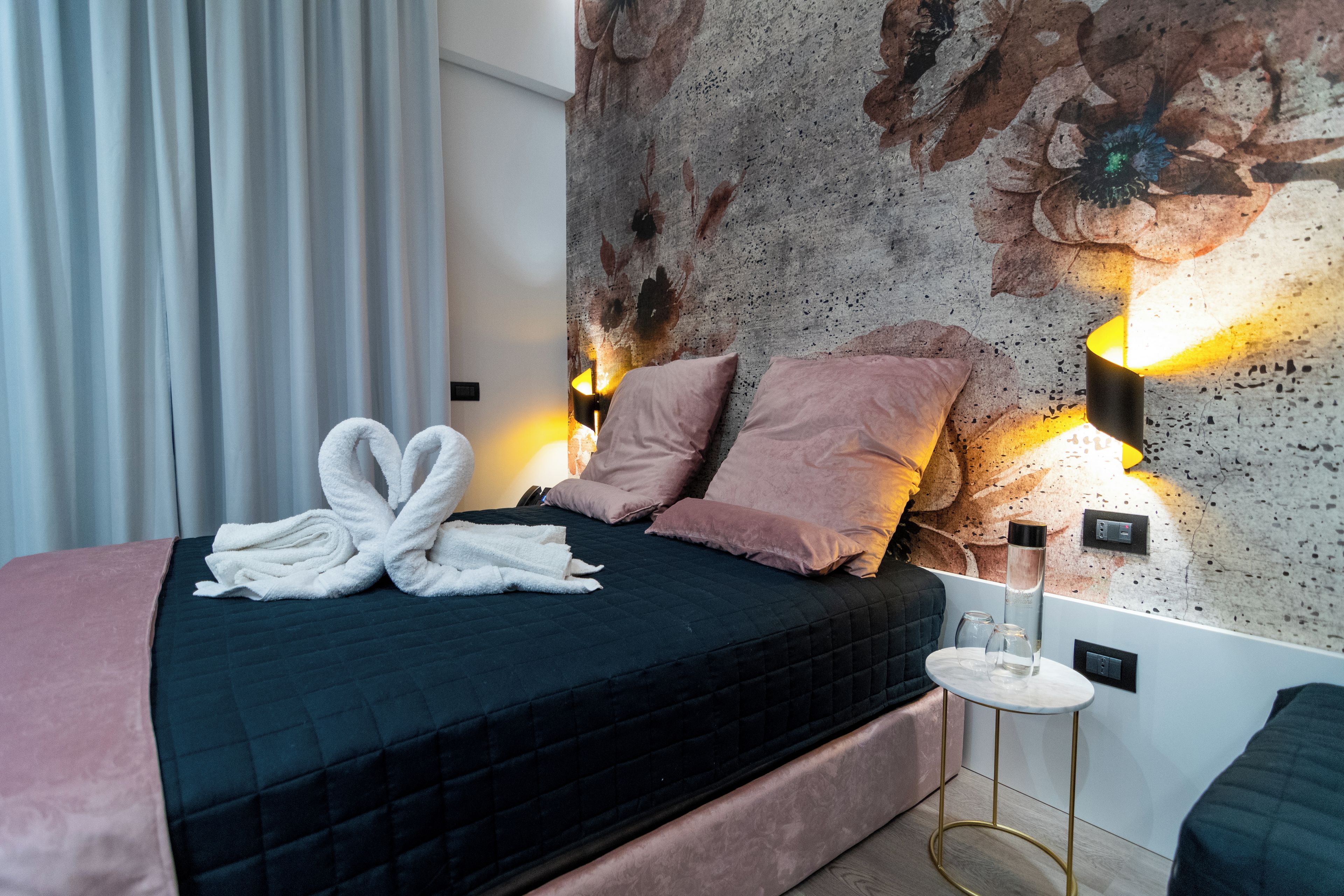 A neatly made bed with a dark quilt, pink pillows, and a pair of white towels folded on top. There is a small round table with a lamp beside the bed.