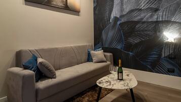 A modern living room with a beige sofa, a small round table with a bottle and glasses, and a large abstract mural on the wall.