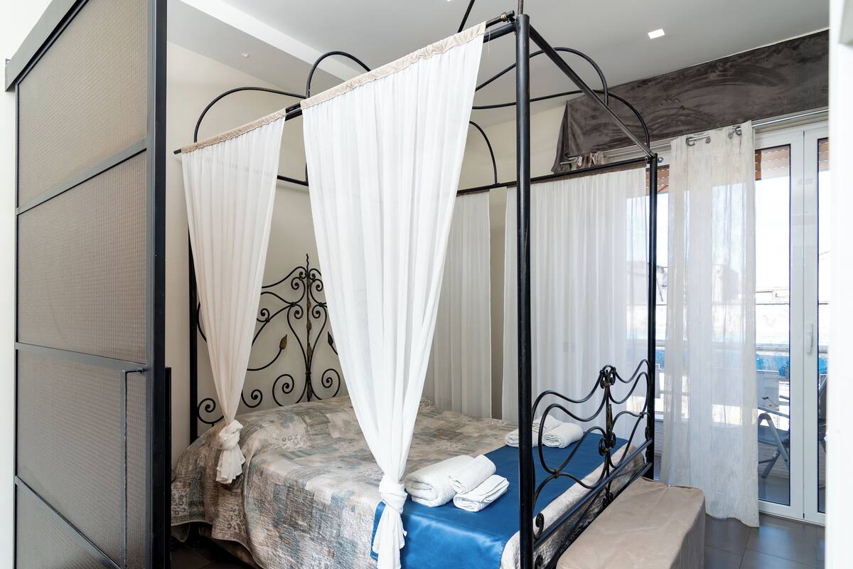 A four-poster canopy bed with white curtains, a blue bedspread, and neatly folded towels on the side.