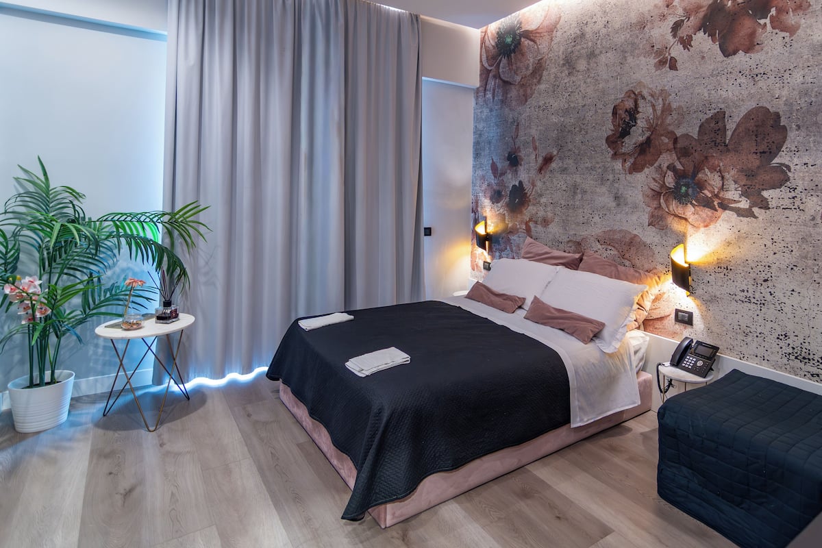 A modern bedroom with a large bed, a bedside table with a lamp, a small side table with a phone, and a wall with a floral mural.
