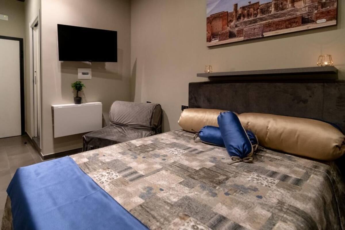 A hotel room with a bed, a television mounted on the wall, a small table with a plant, and a framed picture above the bed.