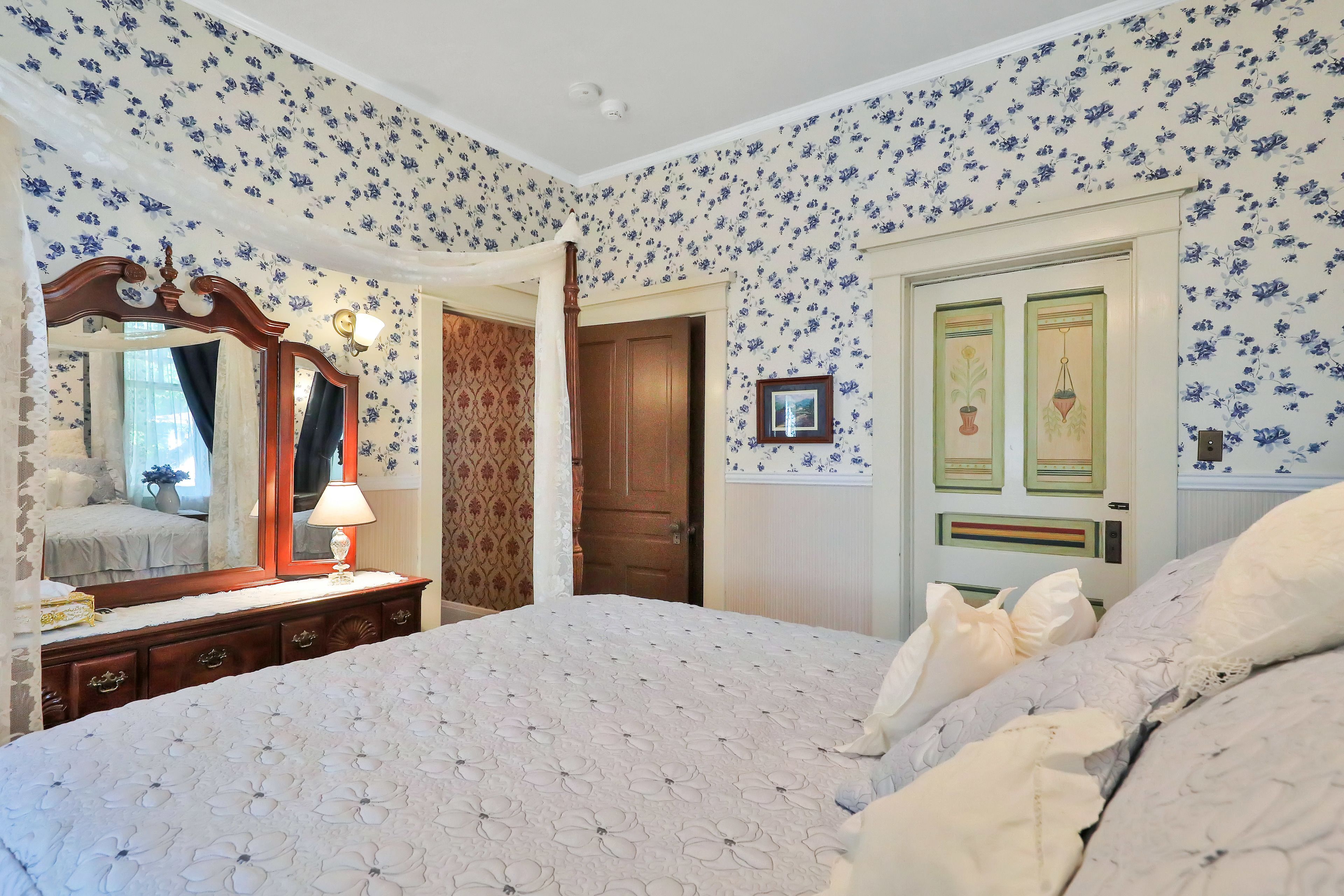 Un dormitorio con papel tapiz floral, una cama con colcha, un aparador con espejo y una puerta con paneles decorativos.
