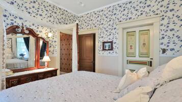 Un dormitorio con papel tapiz floral, una cama con colcha, un aparador con espejo y una puerta con paneles decorativos.