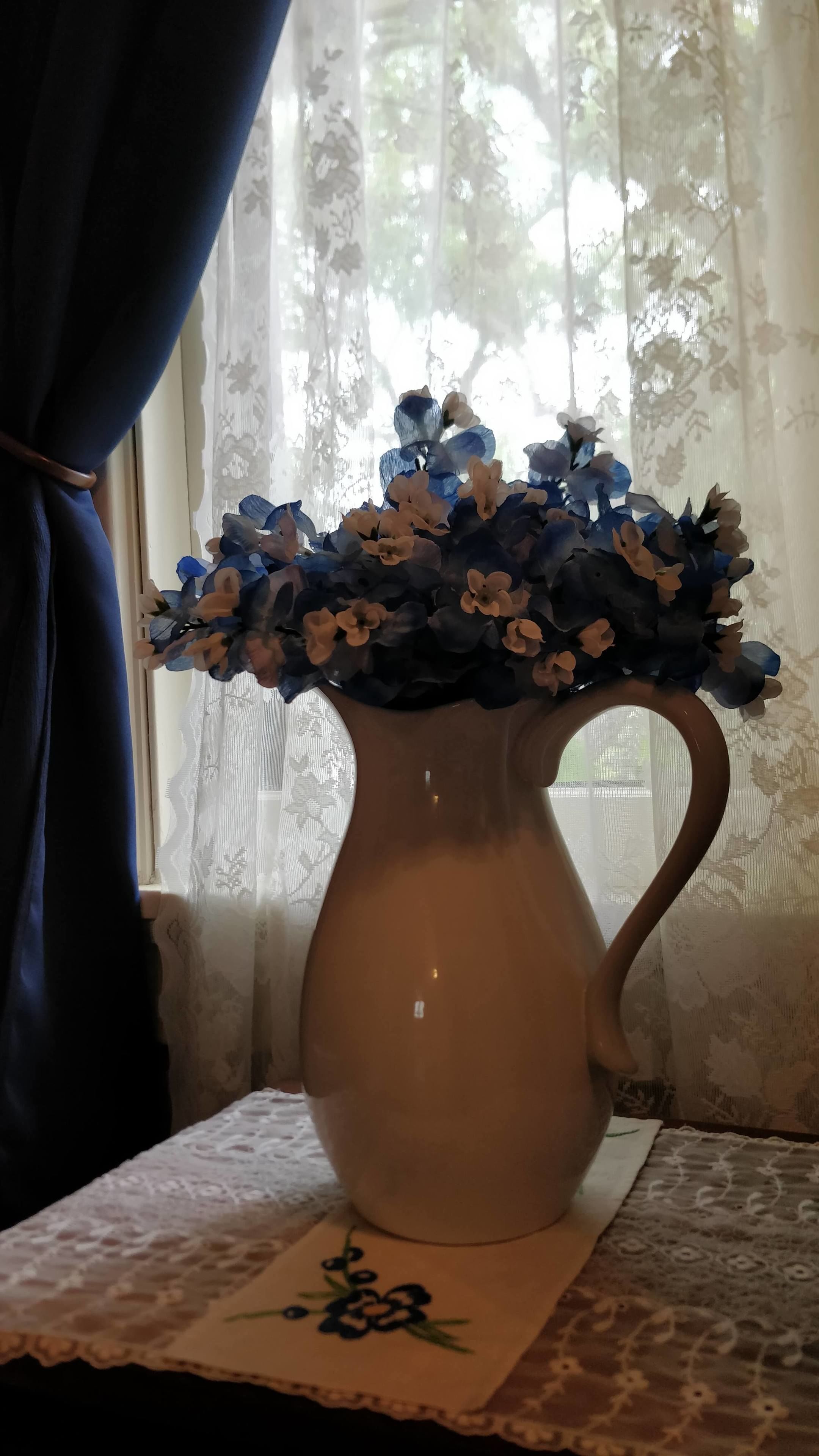 A vase with blue and white flowers on a table near a window with lace curtains.