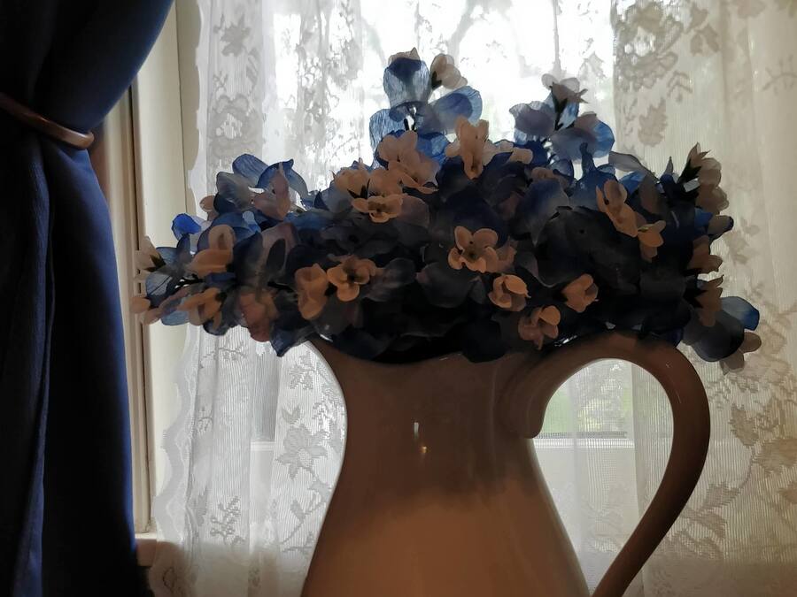 Um vaso com flores azuis e brancas sobre uma mesa perto de uma janela com cortinas de renda.