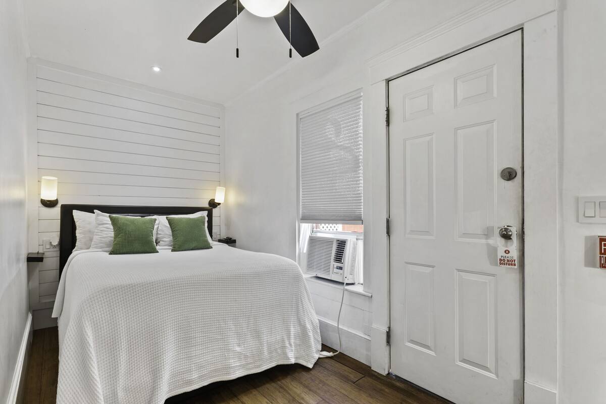Un dormitorio con cama, lámparas de noche, ventilador de techo, ventana con persianas y puerta con letrero.