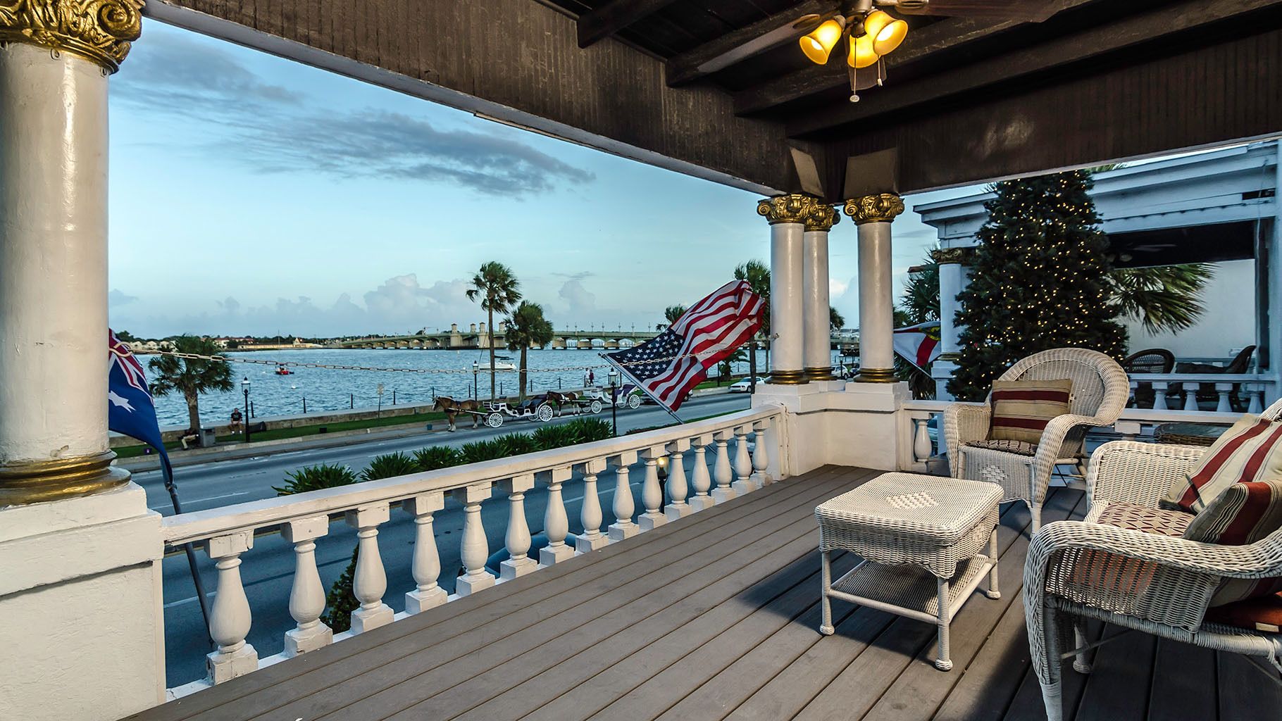 Un balcon avec des fauteuils en osier, un drapeau américain et une vue sur l’eau, avec un bateau et des palmiers.