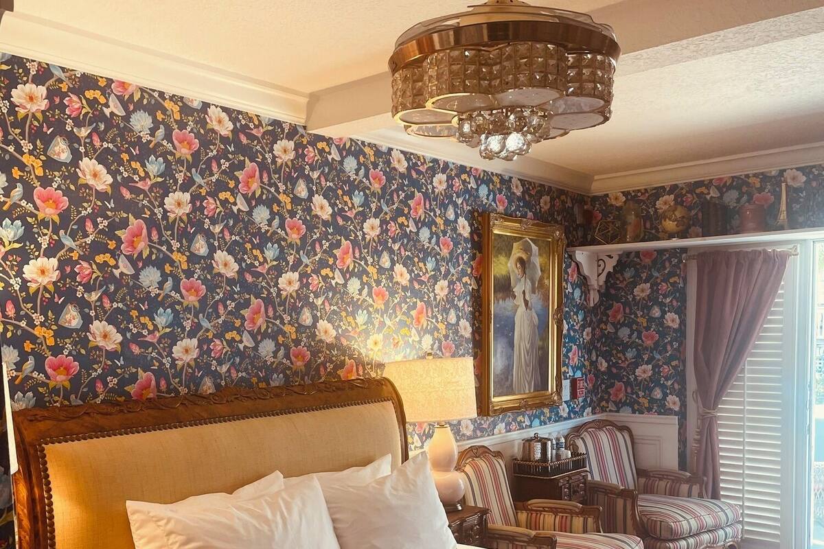 Una habitación de hotel con una cama grande, papel tapiz floral, una araña y un cuadro en la pared.