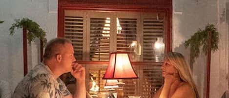 A couple sitting at an outdoor table with string lights overhead, a window with shutters, and potted plants.