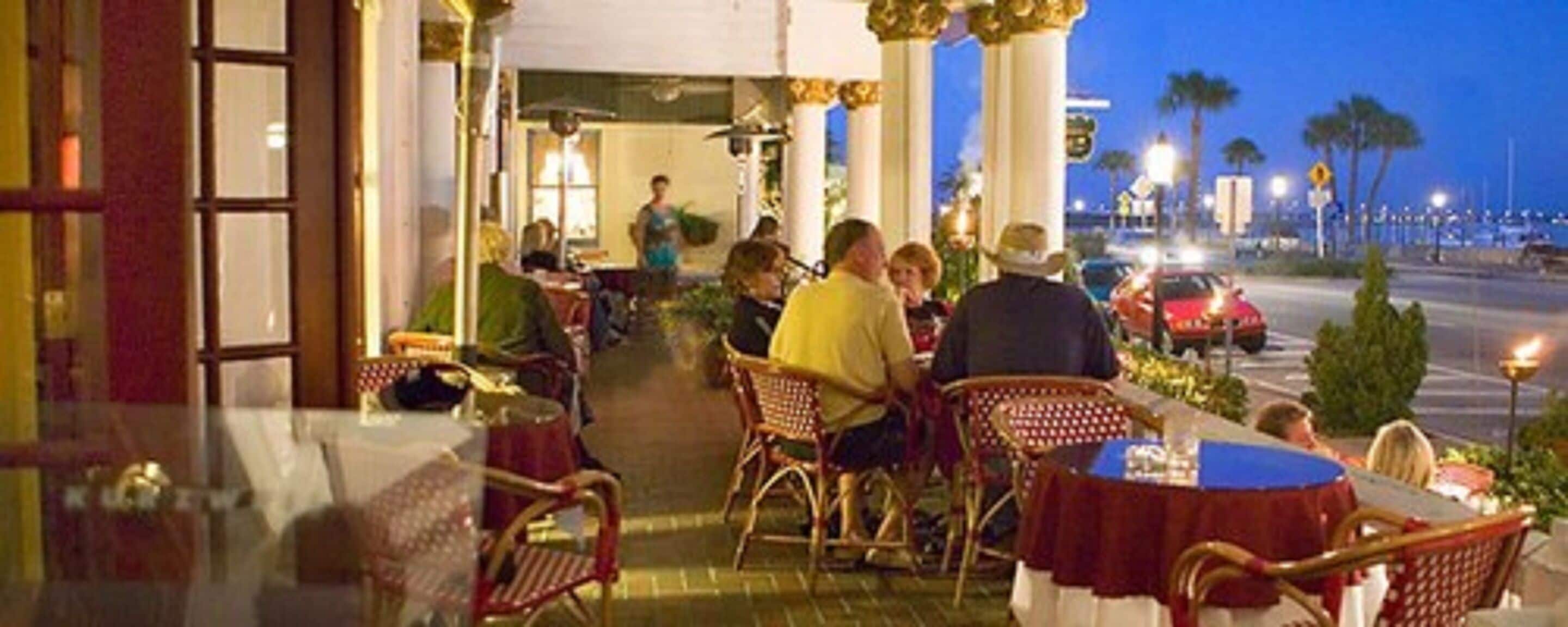 An outdoor dining area with wicker chairs and tables, a pathway leading to the entrance, and a view of the street with parked cars and palm trees.
