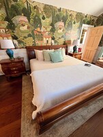 A bedroom with a large bed, wooden headboard, a nightstand with a lamp, a dresser, and a mural on the wall.