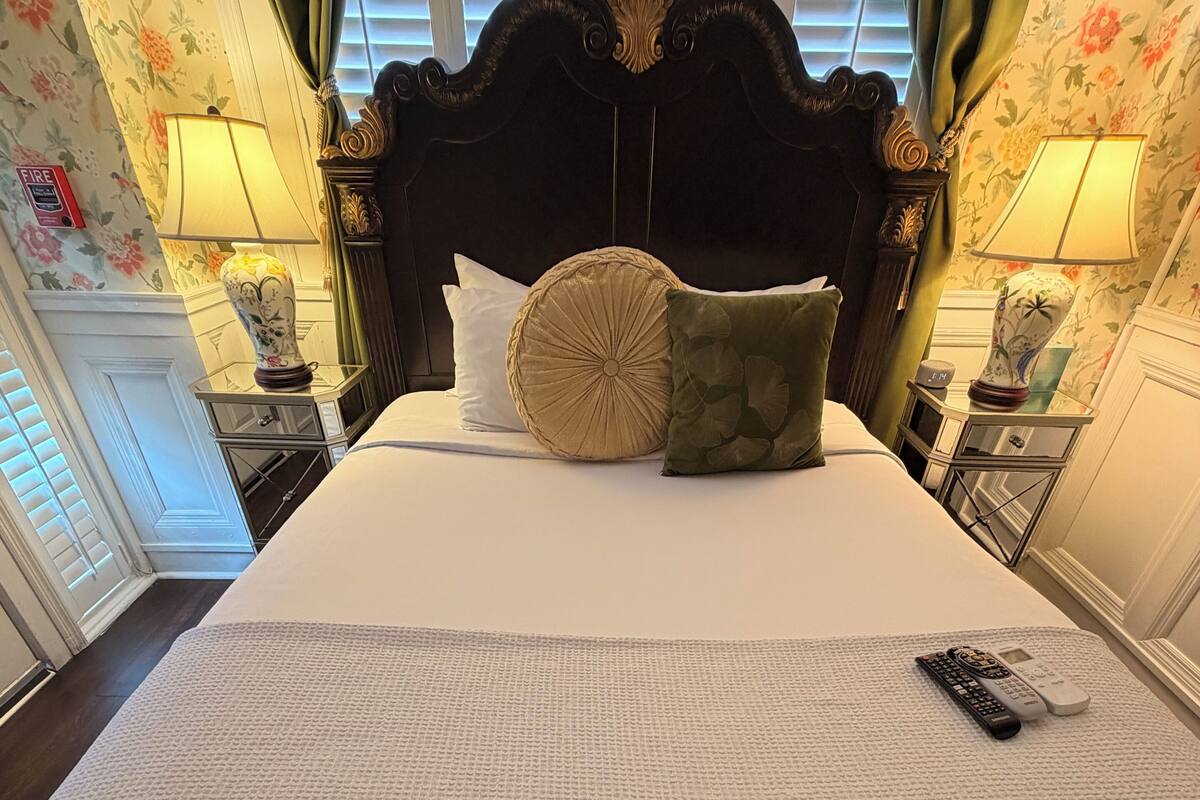 Una cama bien hecha con cabecera, dos lámparas de noche, un control remoto y papel tapiz con motivos florales.