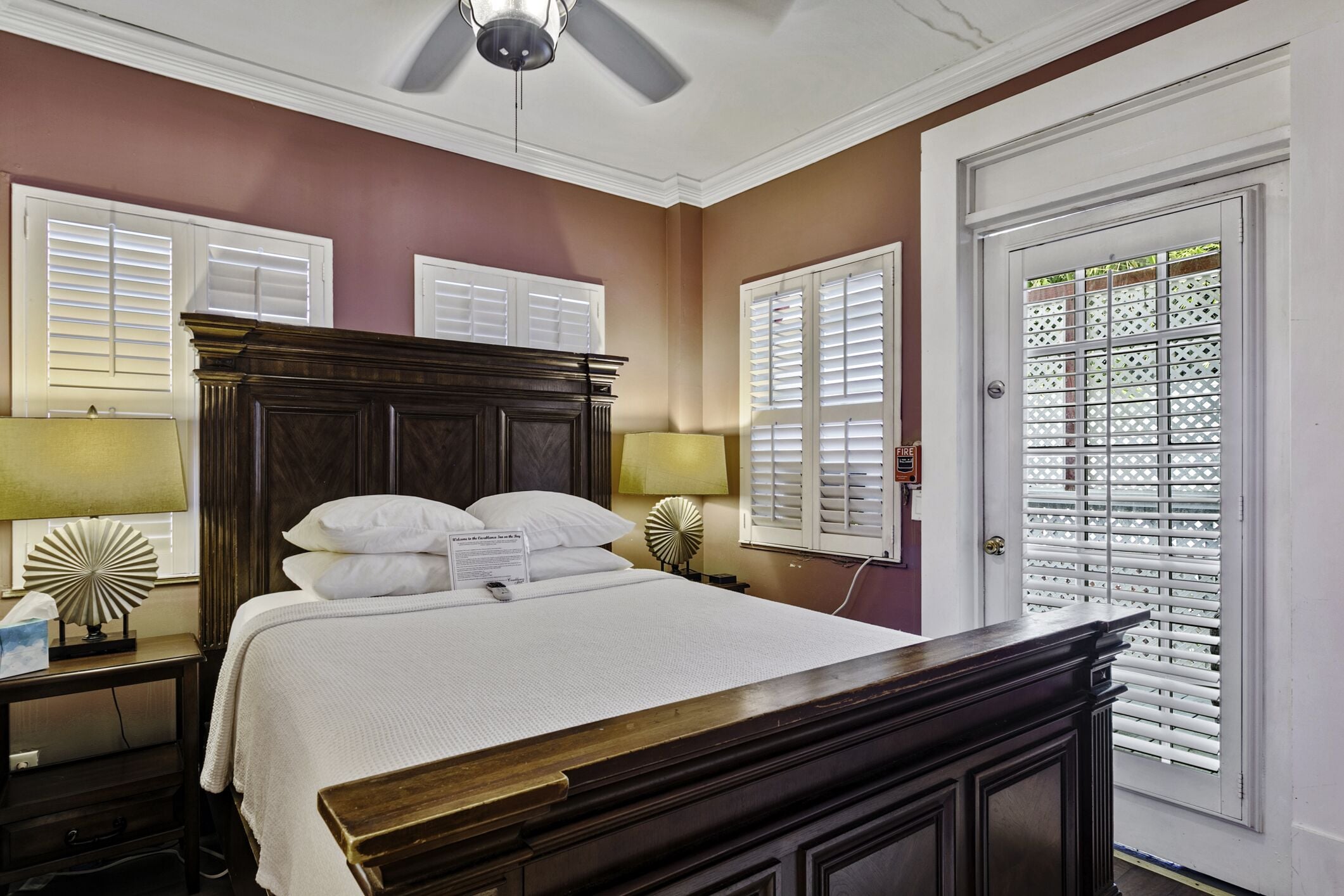 A hotel room with a large bed, two bedside lamps, a ceiling fan, and a door with a decorative window grille.