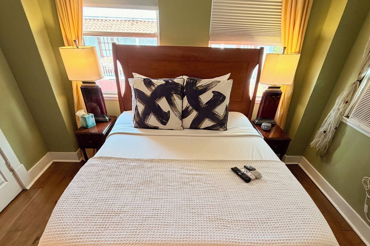 Una cama con colchón blanco texturizado, dos almohadas con estampado blanco y negro, cabecera de madera, dos mesitas de noche con lámparas y suelo de madera.
