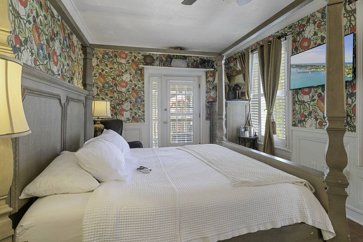 Un dormitorio con una cama grande, papel tapiz floral, un televisor en la pared y una puerta con vistas al exterior.
