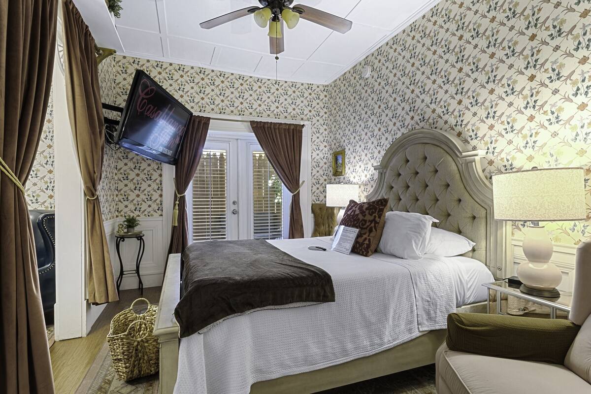 Un dormitorio con cama, televisión, mesita de noche, lámpara y ventana con cortinas.