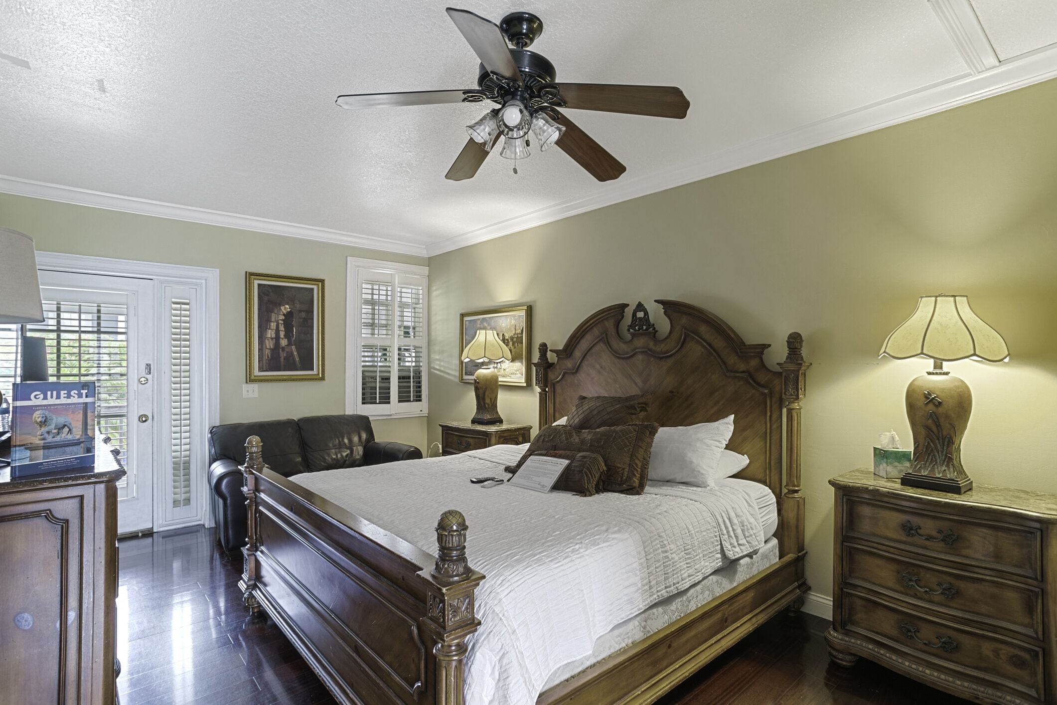 A bedroom with a large bed, a ceiling fan, a nightstand with a lamp, and a dresser.