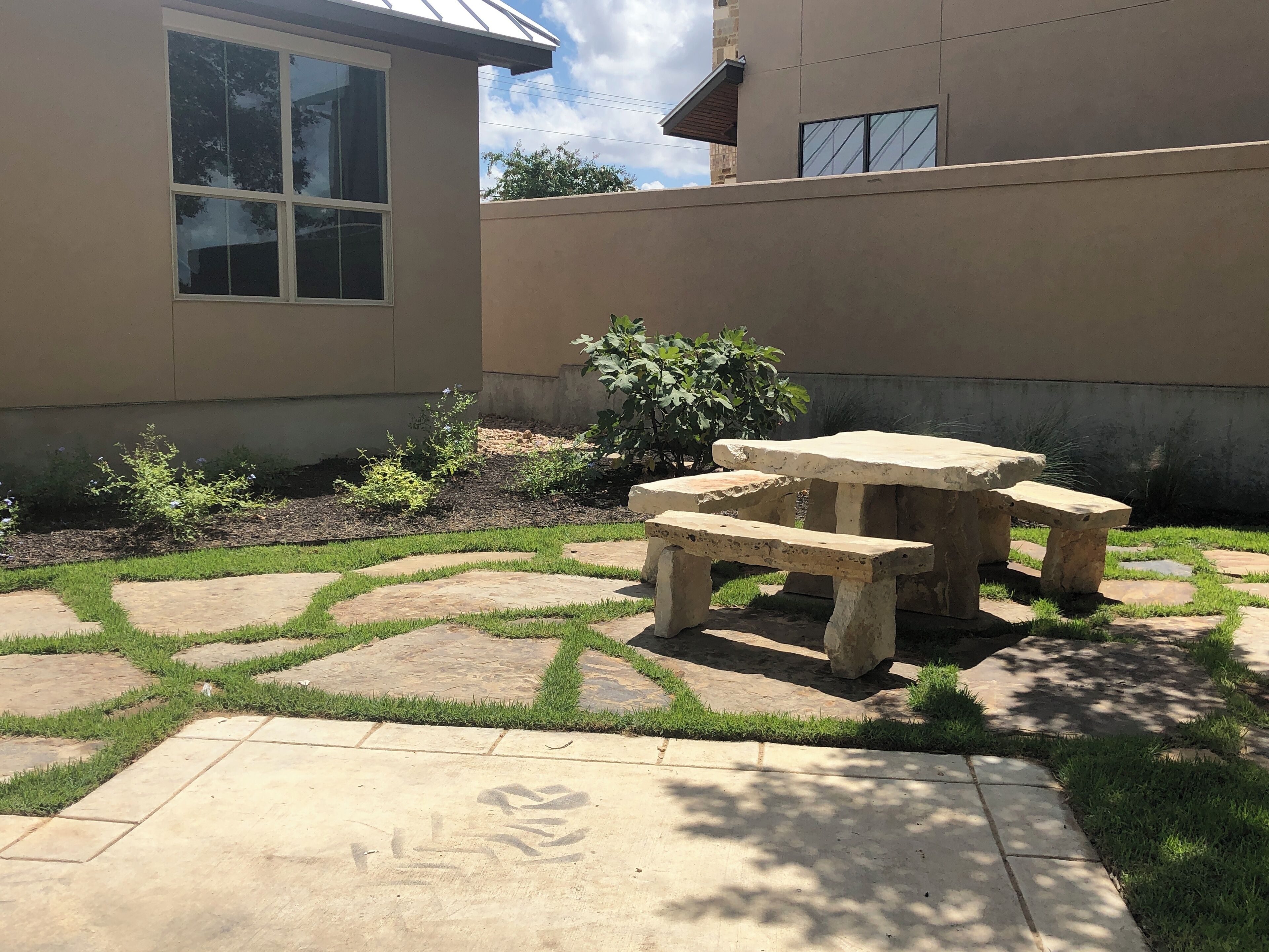 A patio with a stone table and bench, surrounded by a garden with grass and plants.