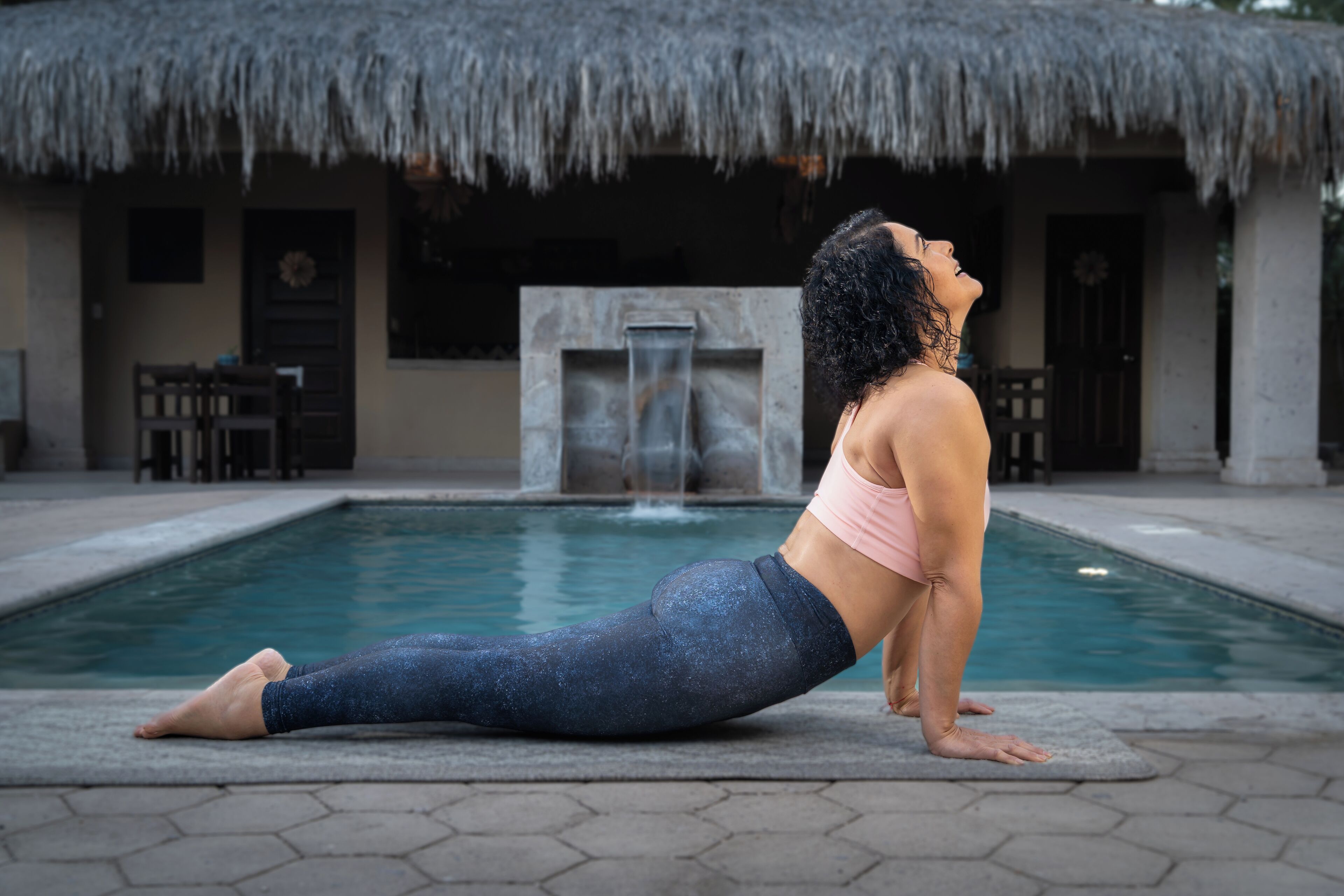 Eine Person führt eine Yoga-Pose auf einer Matte am Pool aus, im Hintergrund ein Gebäude mit einem Reetdach.