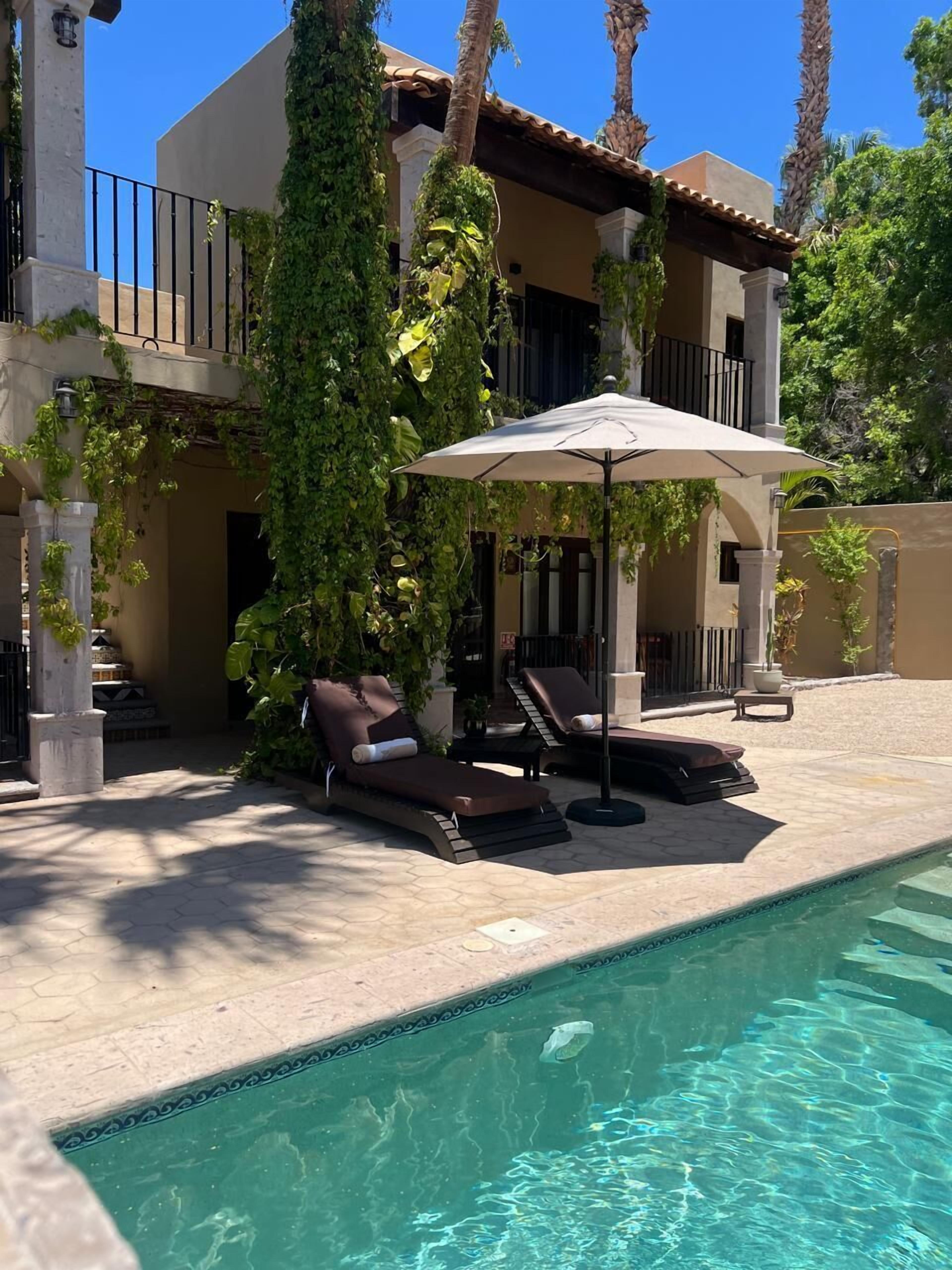 A poolside area with lounge chairs and an umbrella, surrounded by a building with a tiled roof and greenery.