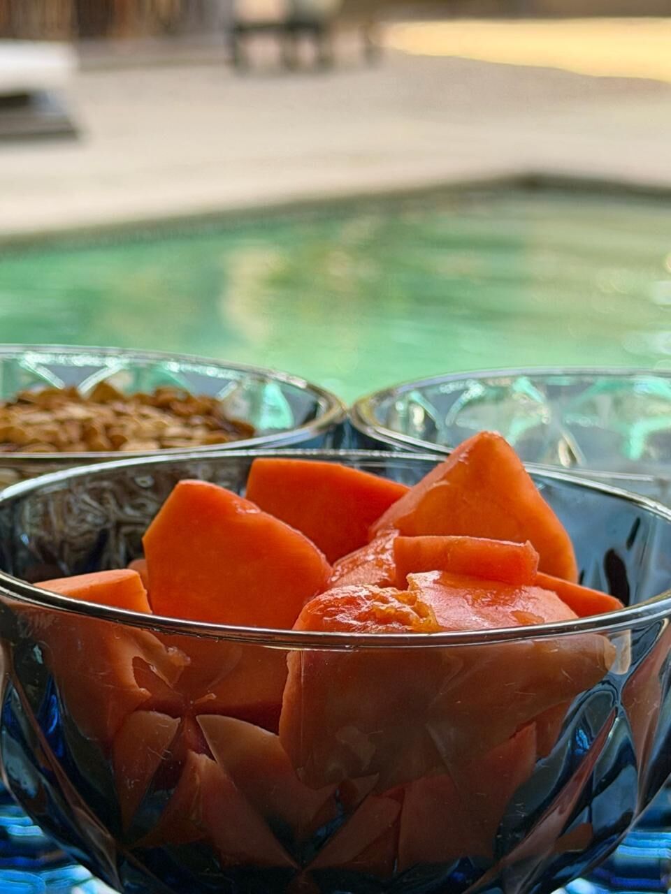 A glass bowl filled with sliced fruit, likely mango, on a table with a pool in the background.