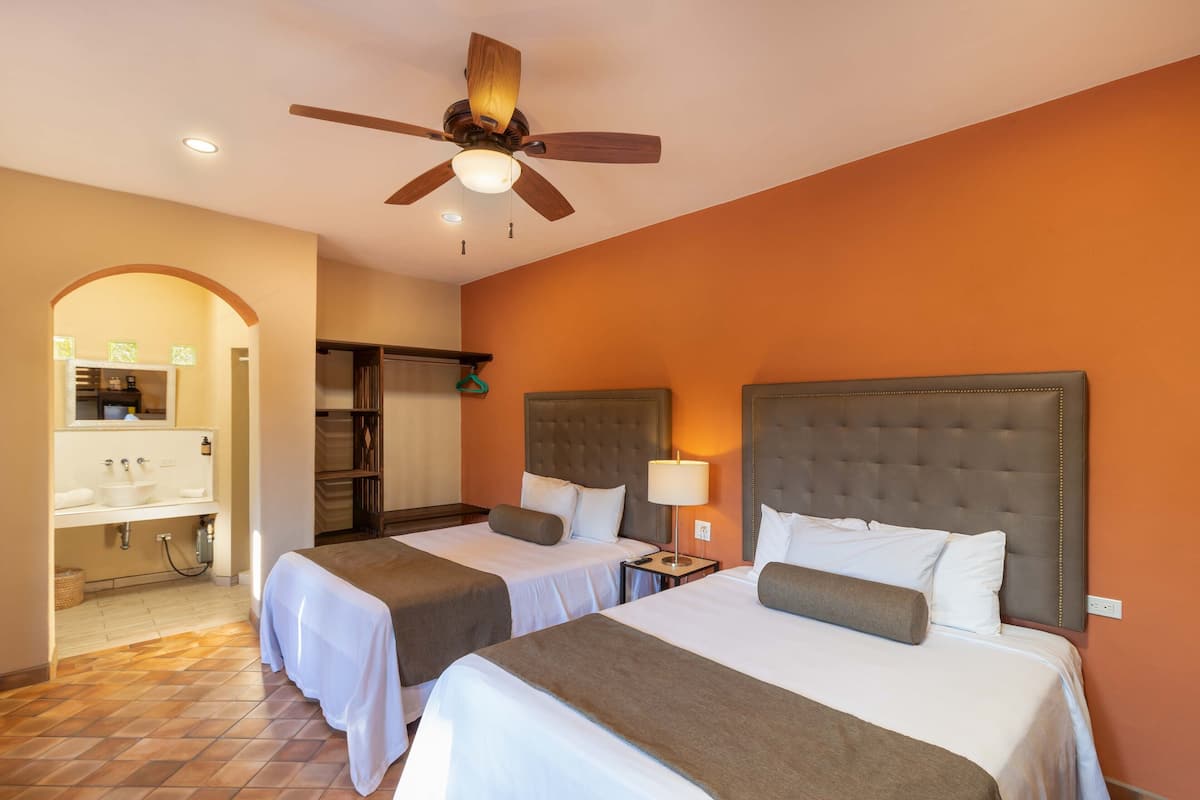 A hotel room with two beds, a ceiling fan, a bathroom with a sink, and a mirror.