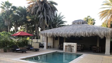 Un área de piscina con sillones, una estructura de techo de paja y palmeras.