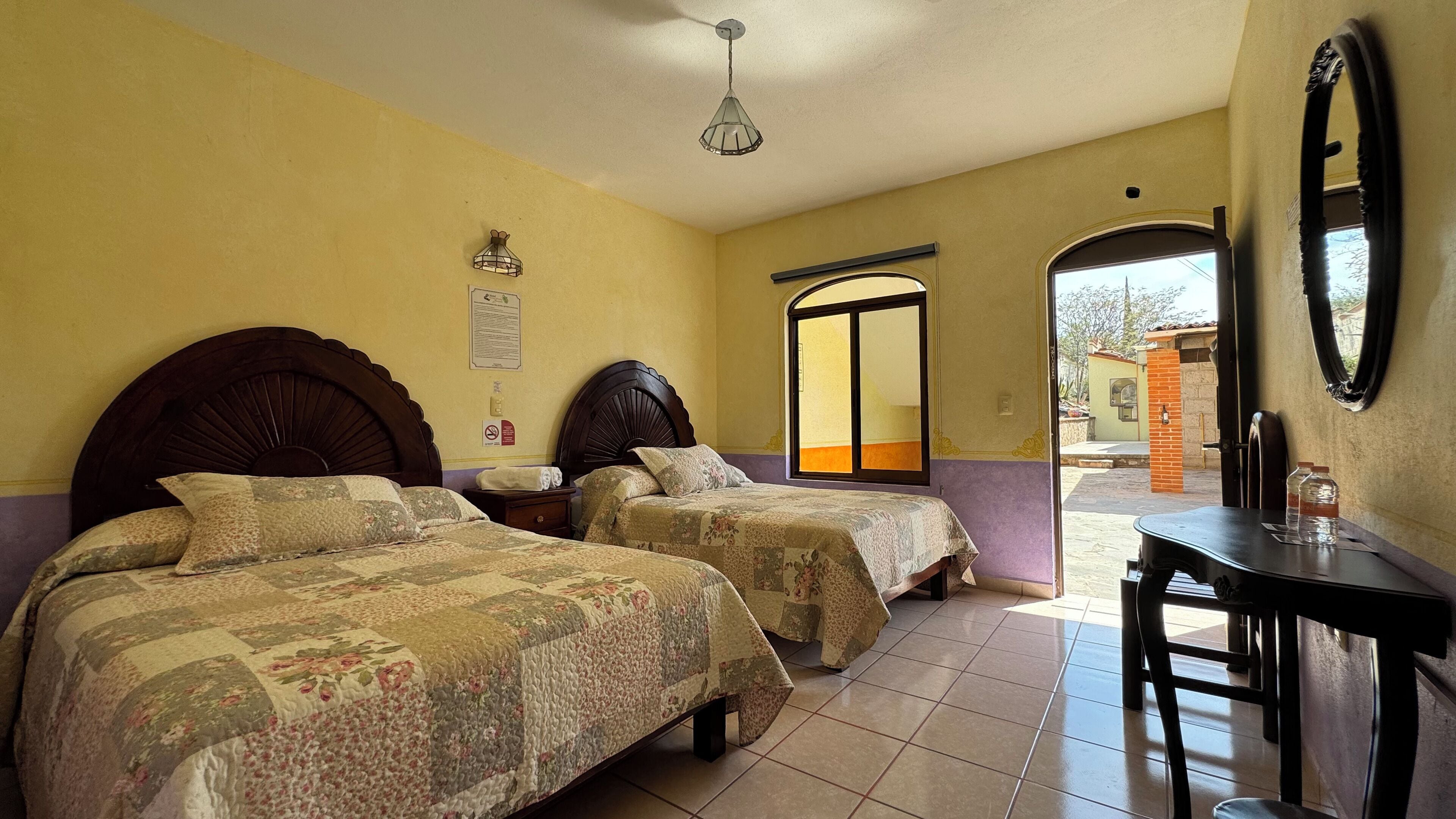 Une chambre d’hôtel avec deux lits, une petite table, un miroir et une fenêtre donnant sur l’extérieur.