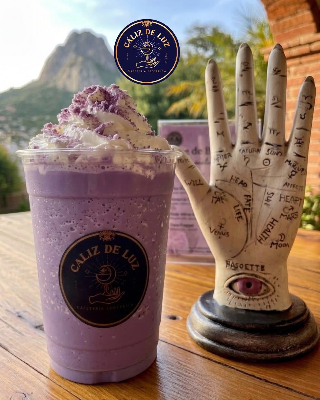 A purple beverage in a branded cup with a logo, next to a decorative hand with zodiac signs.