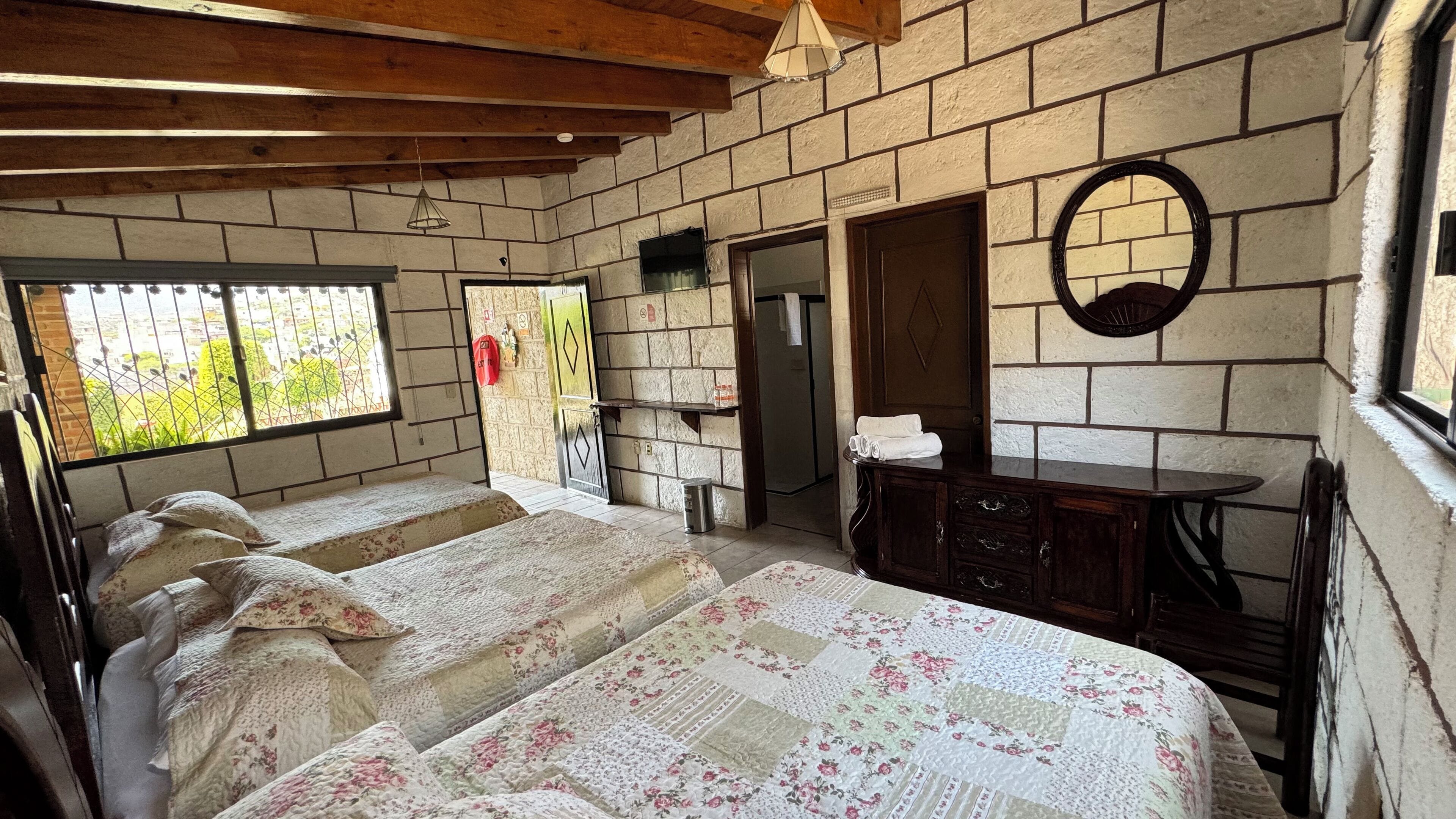 A room with two beds, a small table, a mirror, and a window with a view of greenery.