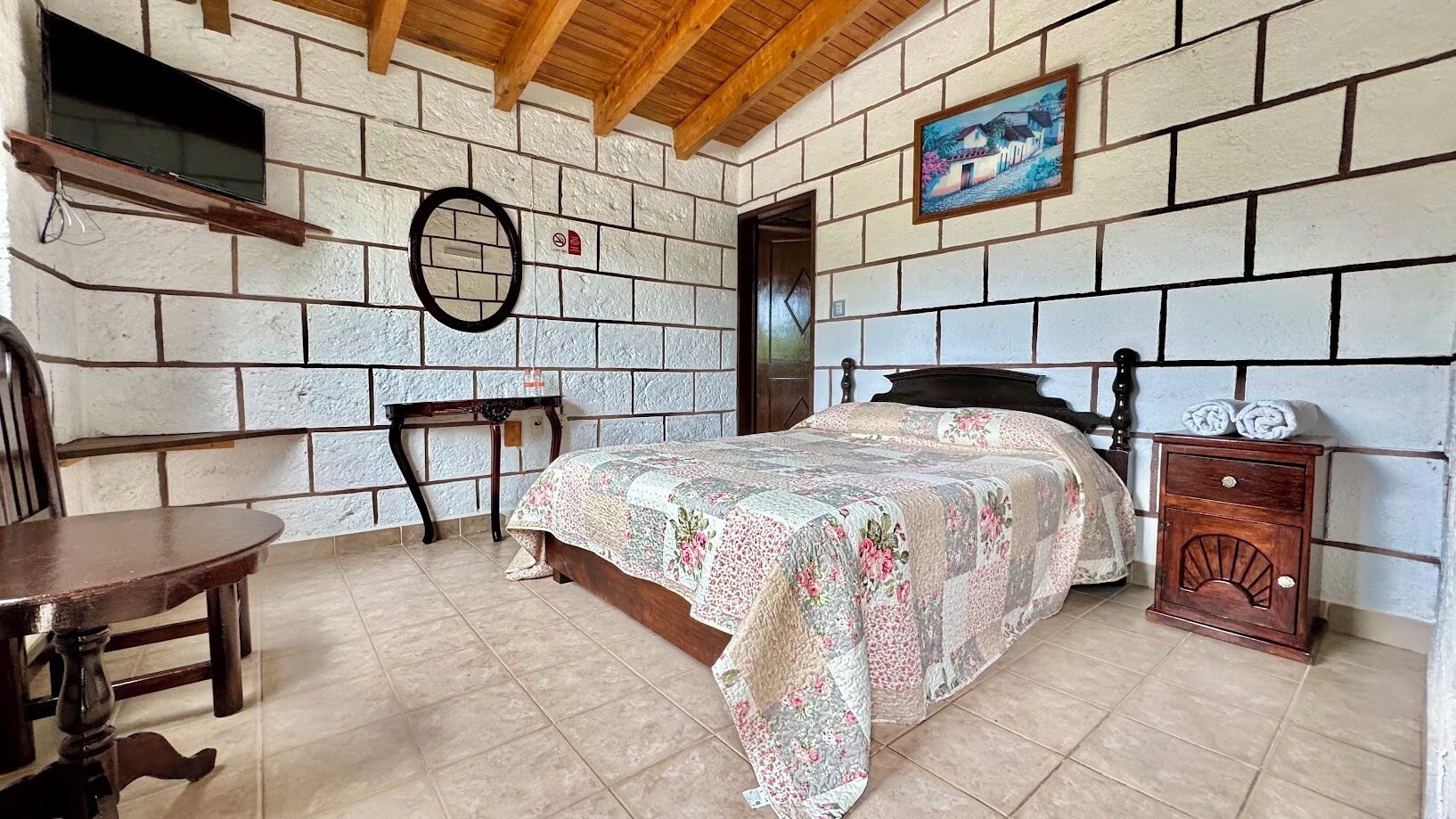 A bedroom with a bed, a wooden nightstand, a small table, and a painting on the wall.