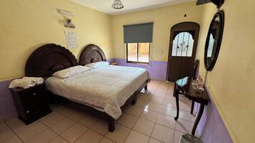 A bedroom with a bed, a nightstand, a chair, a table with bottles, a window, and a door.