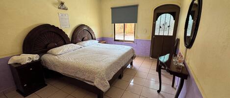 Un dormitorio con una cama, una mesita de noche, una silla, una mesa con botellas, una ventana y una puerta.